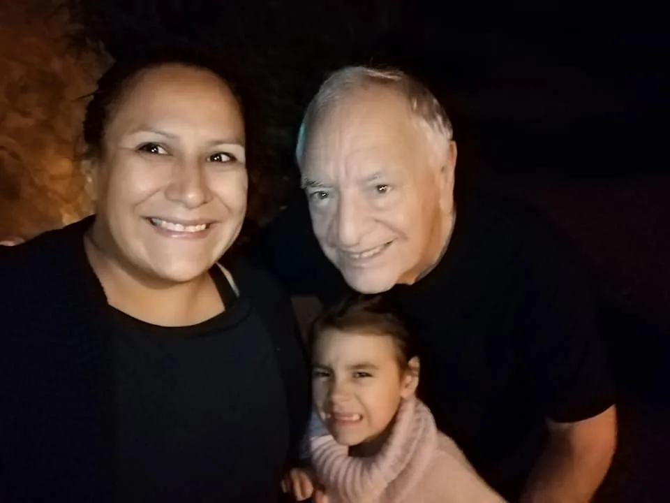 A woman and a white-haired elder smile together at night while a young girl in a pink sweater makes a playful face in front of them.