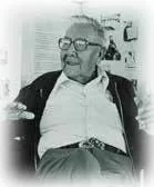 Black-and-white portrait of Chief Mathew King, an elderly man in glasses, a light shirt and dark jacket, seated with one hand raised as he speaks.