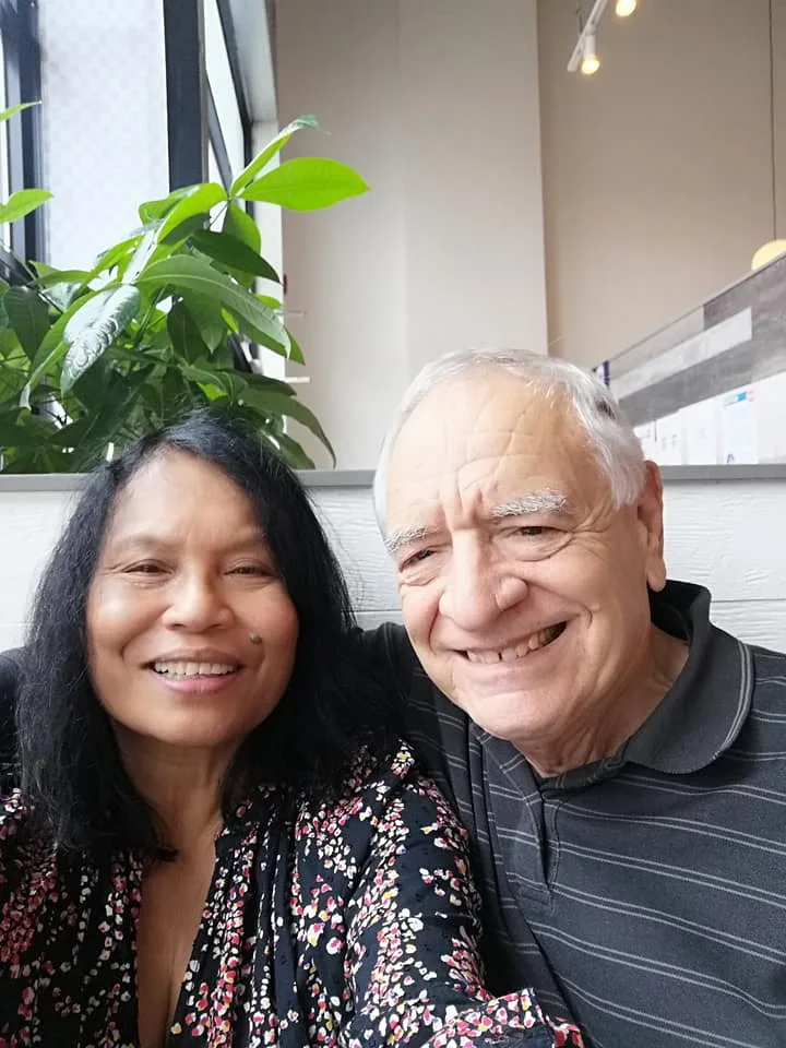 A smiling woman with long dark hair in a floral blouse and a smiling white-haired elder in a striped polo shirt sit together in a bright restaurant beside a leafy plant.