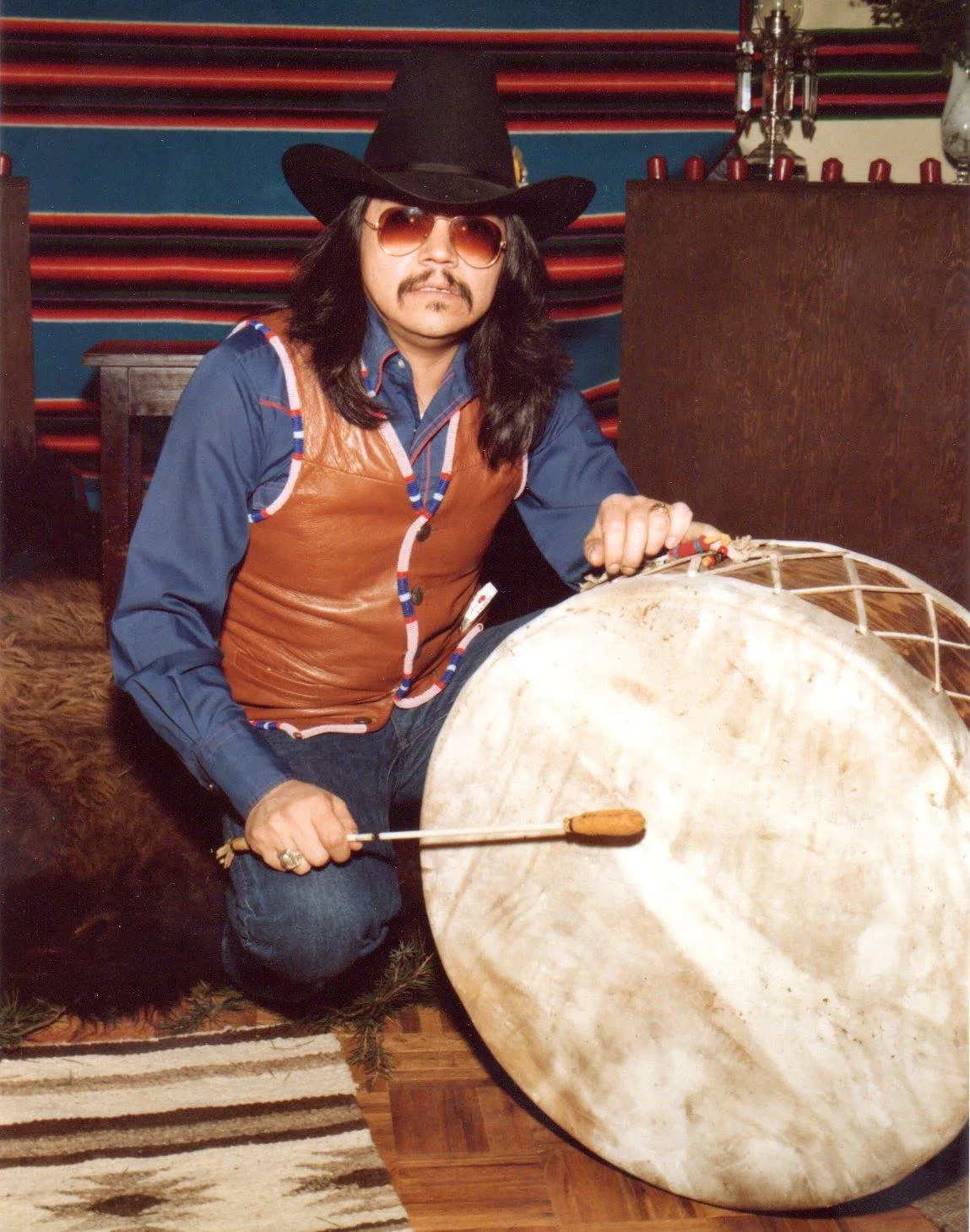 A man in a black cowboy hat, tinted glasses and beaded leather vest kneels beside a large hand drum, holding a drumstick, before a striped blanket.