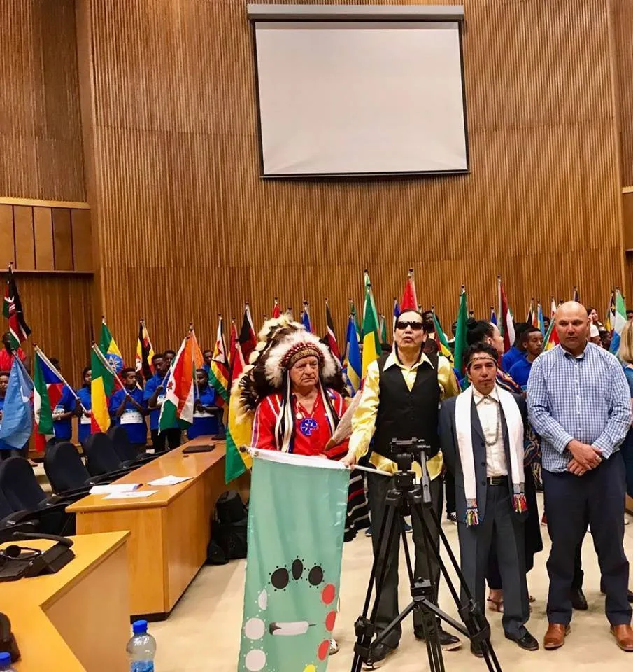 A man in a feather headdress and red regalia holds a green Indigenous Unity Flag beside others in a hall at the African Union, with young people holding national flags behind them.