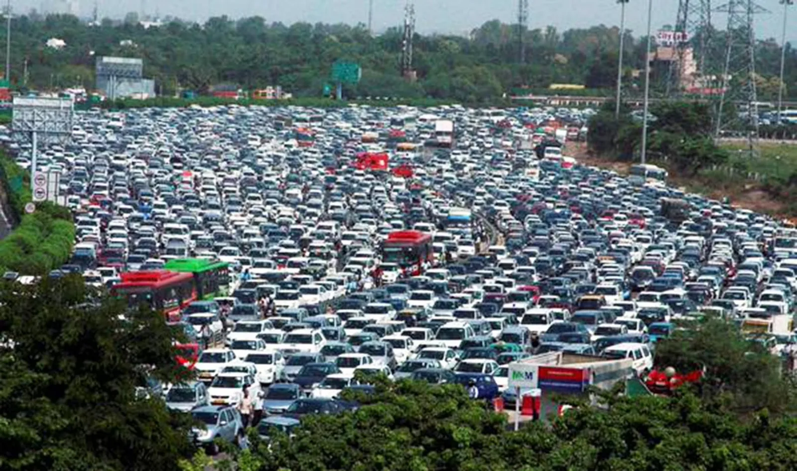 A massive traffic jam of cars and red and green buses packed across a wide multi-lane highway, bordered by trees and power line towers.