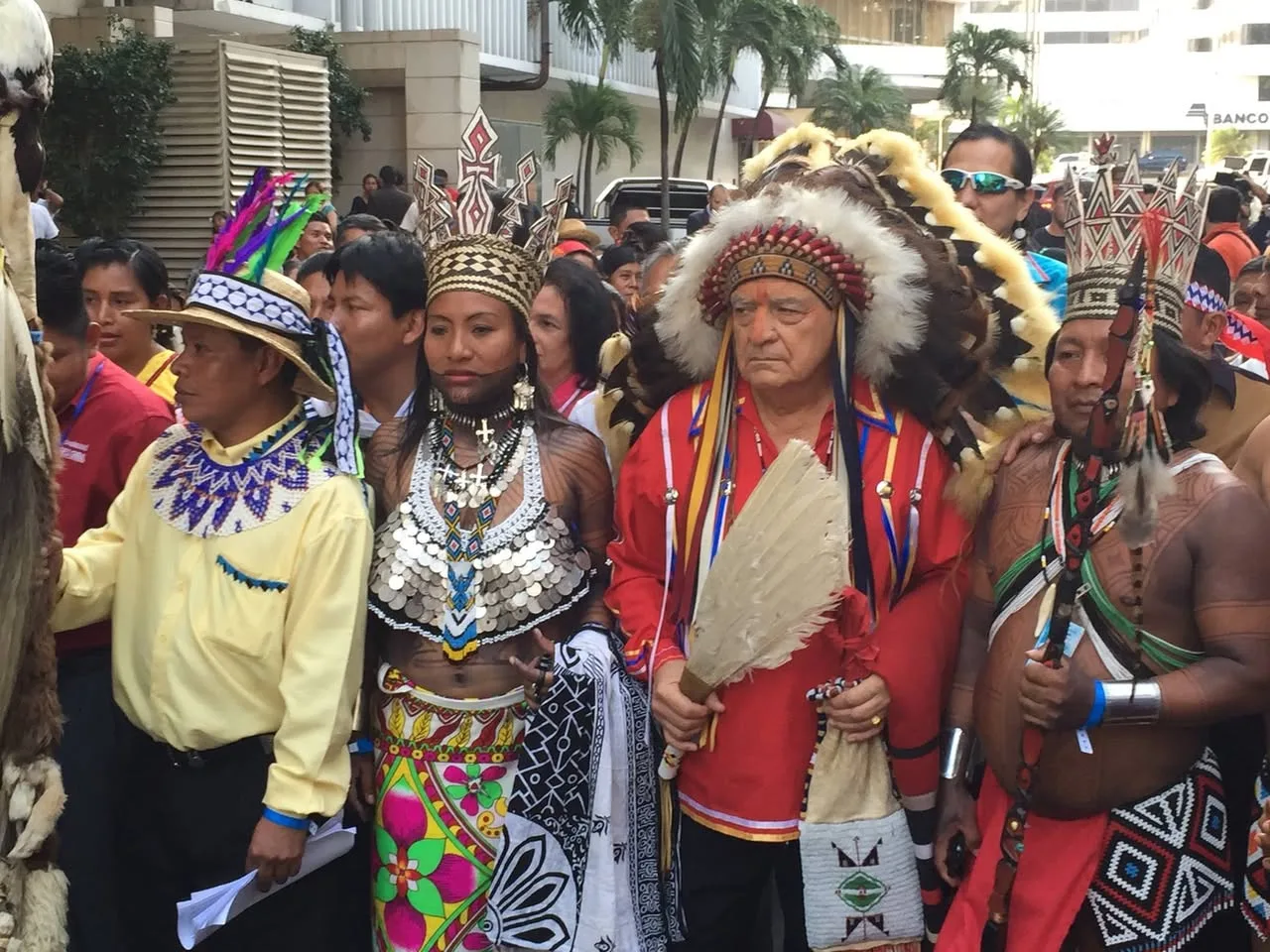 Indigenous delegates from across the Americas in traditional regalia, including an elder in a feather headdress holding a feather fan, gather on a city street.