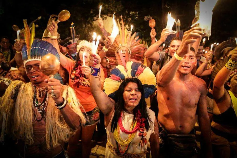 A crowd of Indigenous people at night, many in feather headdresses and beaded necklaces, raise lit candles and rattles as they chant together.