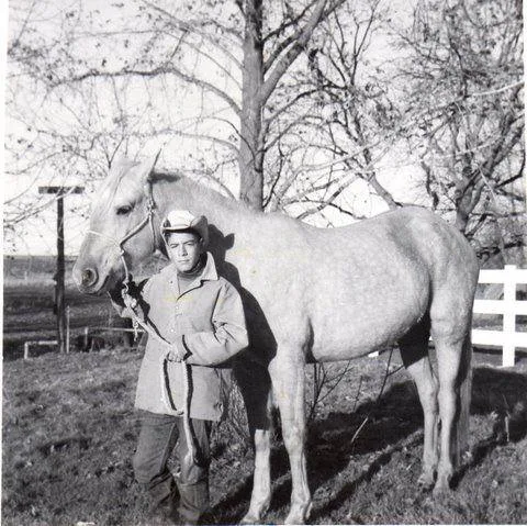 Black-and-white photo of a young boy in a cowboy hat and jacket holding the lead rope of a light gray horse, with bare trees and a white fence behind them.