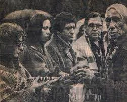 Black-and-white newspaper photo of a group standing close together outdoors, including women with long hair and men in coats, with an elder in a hat at right holding a pipe.
