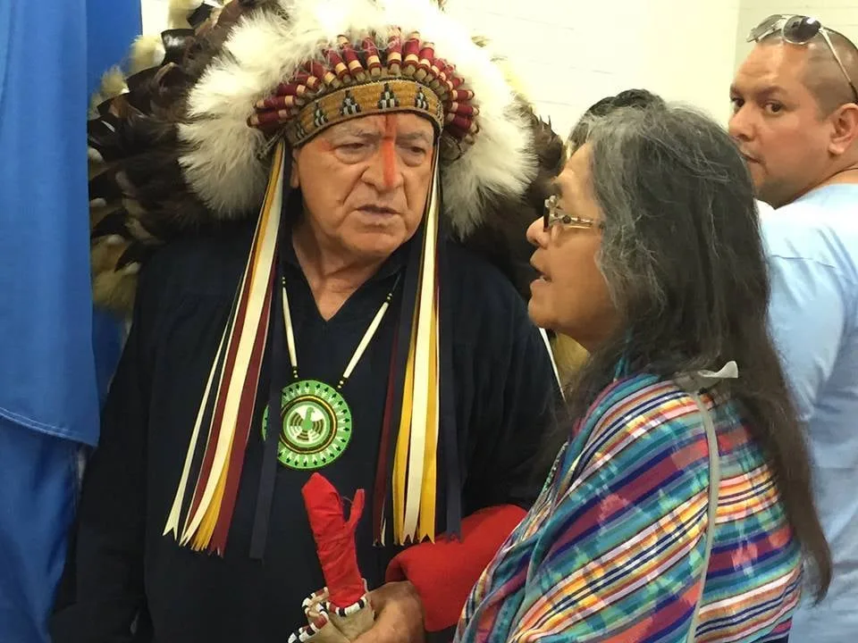 An elder in a feathered headdress with a red-painted forehead and beaded medallion speaks with Mona Polacca, a gray-haired woman in glasses and a striped shawl, indoors.