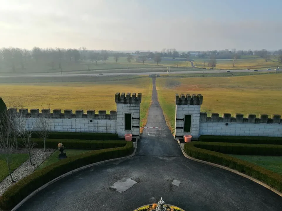 Elevated view of a driveway passing between two crenellated stone gate towers toward golden fields and a distant highway.