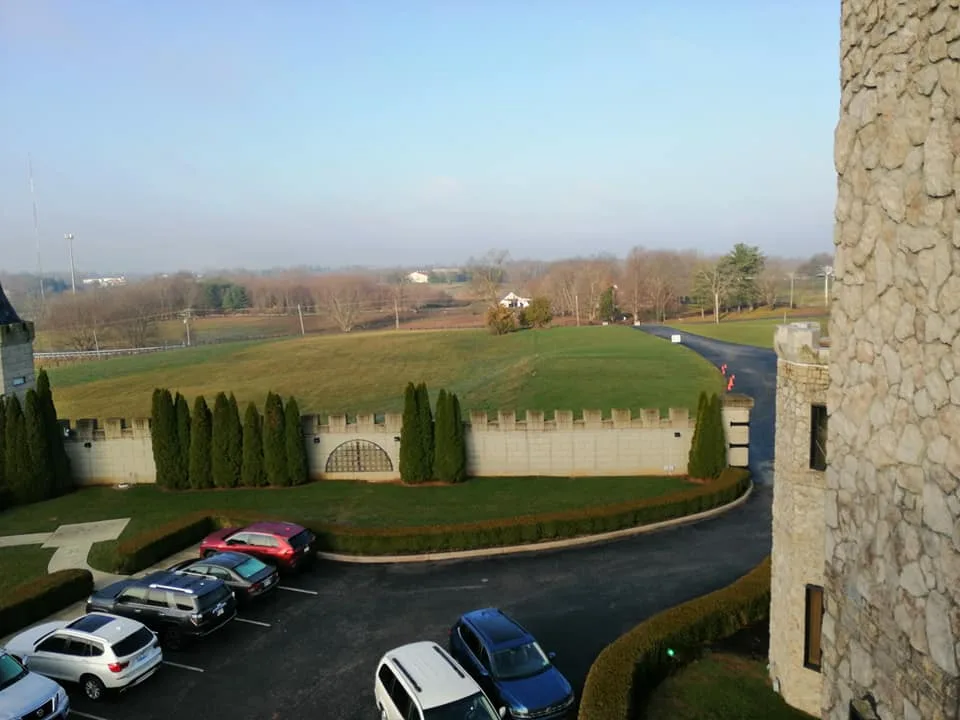View from a stone tower over parked cars, a crenellated wall lined with cypress trees, and green hills under a hazy sky.