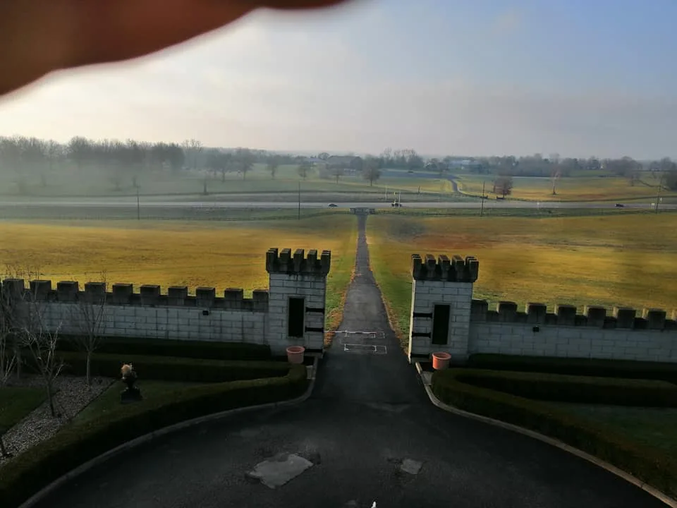 Driveway leading through crenellated gate towers to misty golden fields, with a finger partly covering the top of the frame.