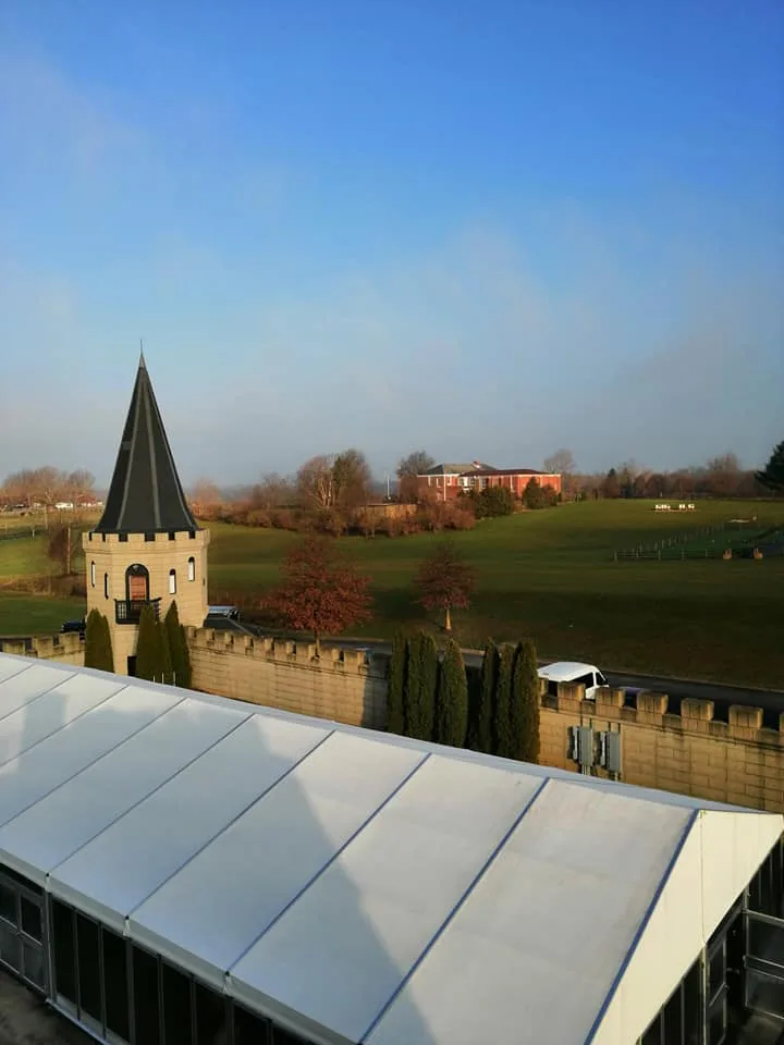 A white tent roof below a castle wall and spired turret, with green fields and a red brick building under blue sky.
