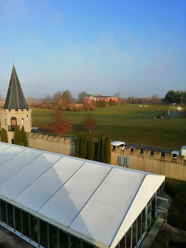 Castle turret with a pointed spire beside a crenellated wall and white tent, overlooking green fields and a brick house.
