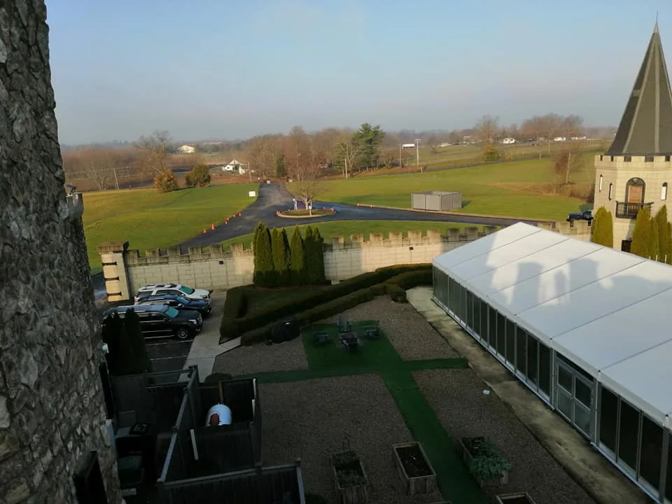 View from beside a stone tower over a courtyard, white tent, crenellated wall, winding driveway and open countryside.