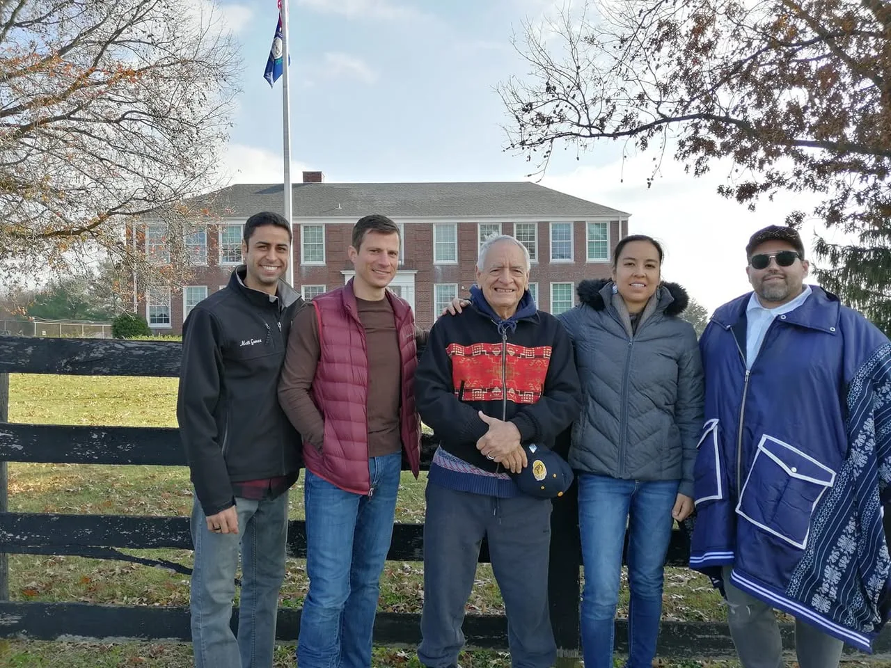 Five people stand smiling by a black wooden fence in front of a two-story red brick building with a flagpole, under autumn trees. An elder in a patterned jacket stands in the centre.
