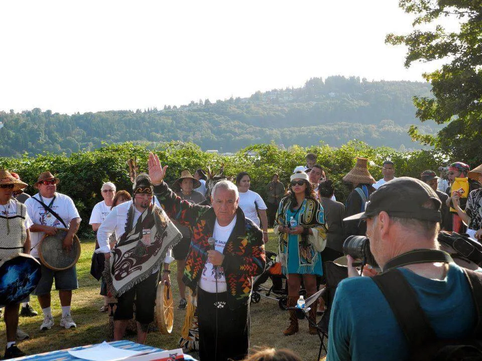 An elder in a patterned blanket raises his hand while speaking into a microphone at an outdoor gathering, surrounded by drummers in cedar hats and regalia, with wooded hills behind.