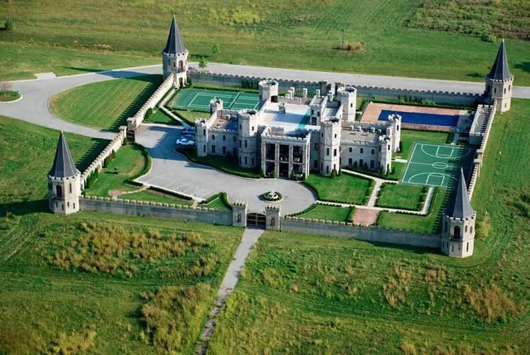 Aerial view of a large stone castle with pointed corner towers and crenellated walls, surrounded by green fields, with a tennis court and basketball court inside.