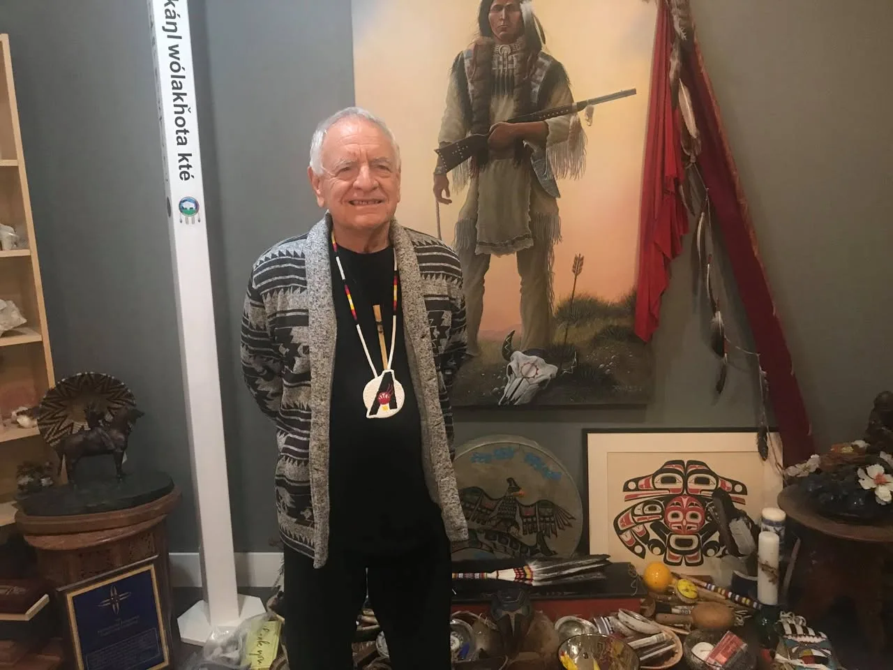 A smiling white-haired elder in a patterned cardigan and beaded medallion stands before a painting of a Plains warrior, a peace pole, feathers, Northwest Coast art and sacred items.