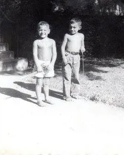 Black-and-white photo of two young shirtless boys standing barefoot on a path, one in shorts and one in long pants, holding thin rods like spears.