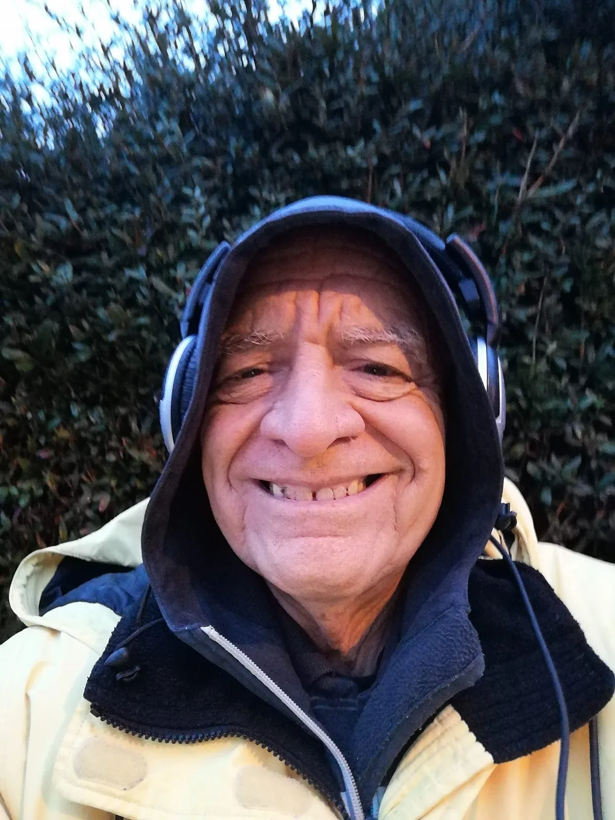 A smiling older man in a dark hood and headphones, wearing a pale yellow jacket, takes a selfie in front of a tall green hedge.