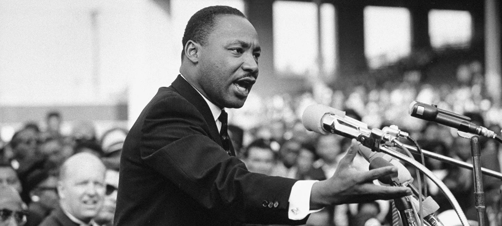 Black-and-white photograph of Dr. Martin Luther King Jr. in a dark suit speaking passionately at a cluster of microphones, one hand outstretched, before a large crowd.