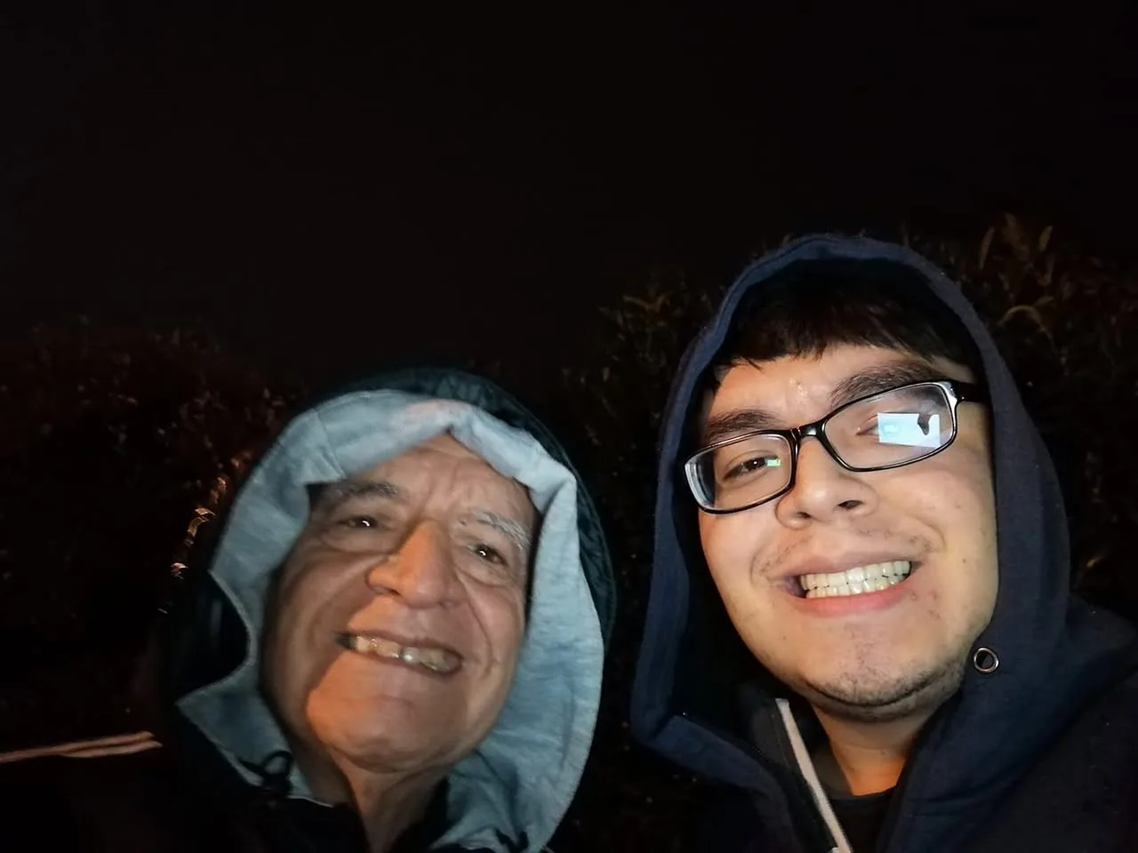 Nighttime selfie of two smiling men in hooded sweatshirts, an older man in a grey-lined hood and a younger man in glasses and a navy hood, with dark foliage behind.