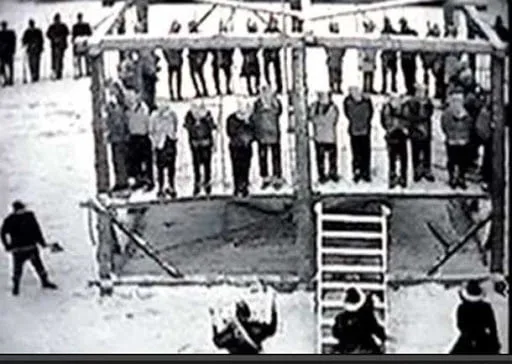 Black-and-white archival image of a large wooden gallows in the snow, with rows of Dakota men standing on the platform as soldiers and onlookers gather below.