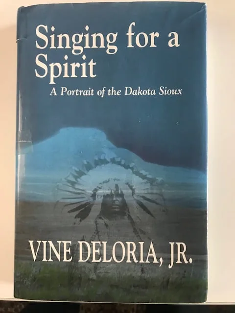 Book cover of "Singing for a Spirit: A Portrait of the Dakota Sioux" by Vine Deloria, Jr., showing a faint image of a man in a feather headdress over a blue butte and prairie.