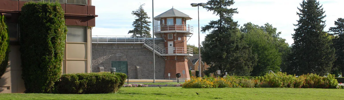 A brick guard tower beside a stone prison wall at the Washington State Penitentiary, with a green lawn, flower beds and evergreen trees in front.
