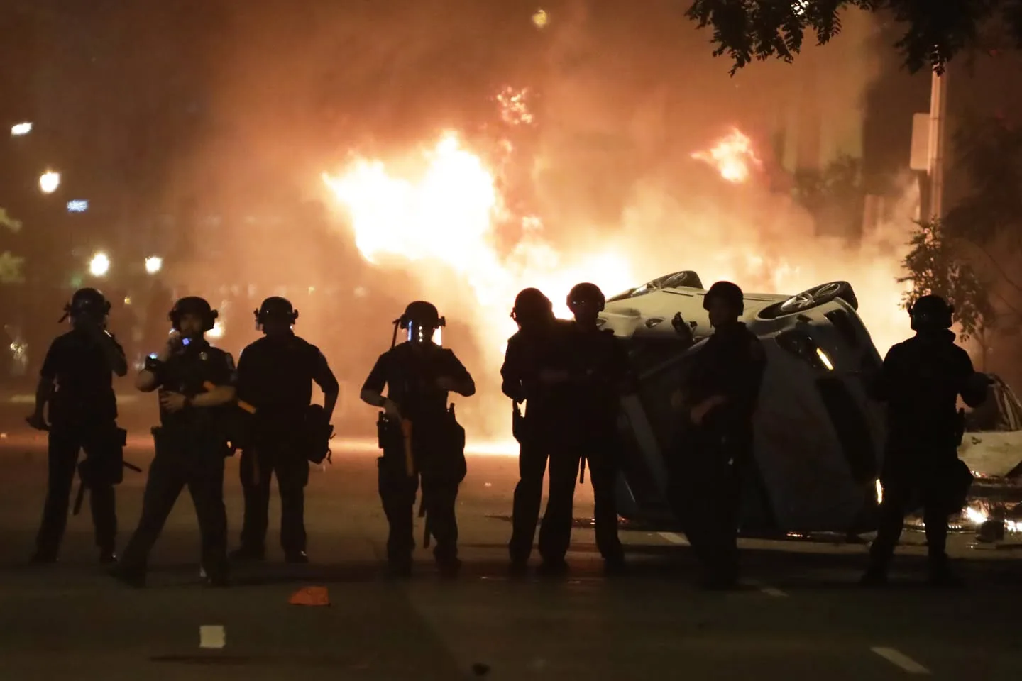 Silhouetted police officers in riot helmets stand in a line on a night street, with an overturned car and large fires blazing behind them.