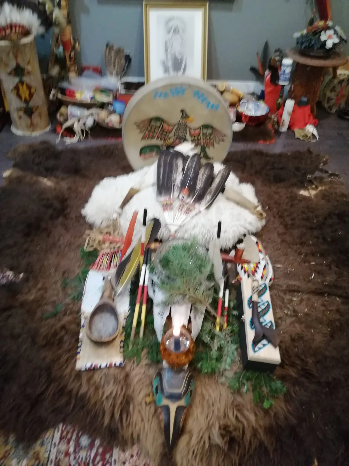 A ceremonial altar on a buffalo robe: a drum painted with a thunderbird, eagle feathers on white fur, a sacred pipe, cedar boughs, a lit candle and a carved bird mask.