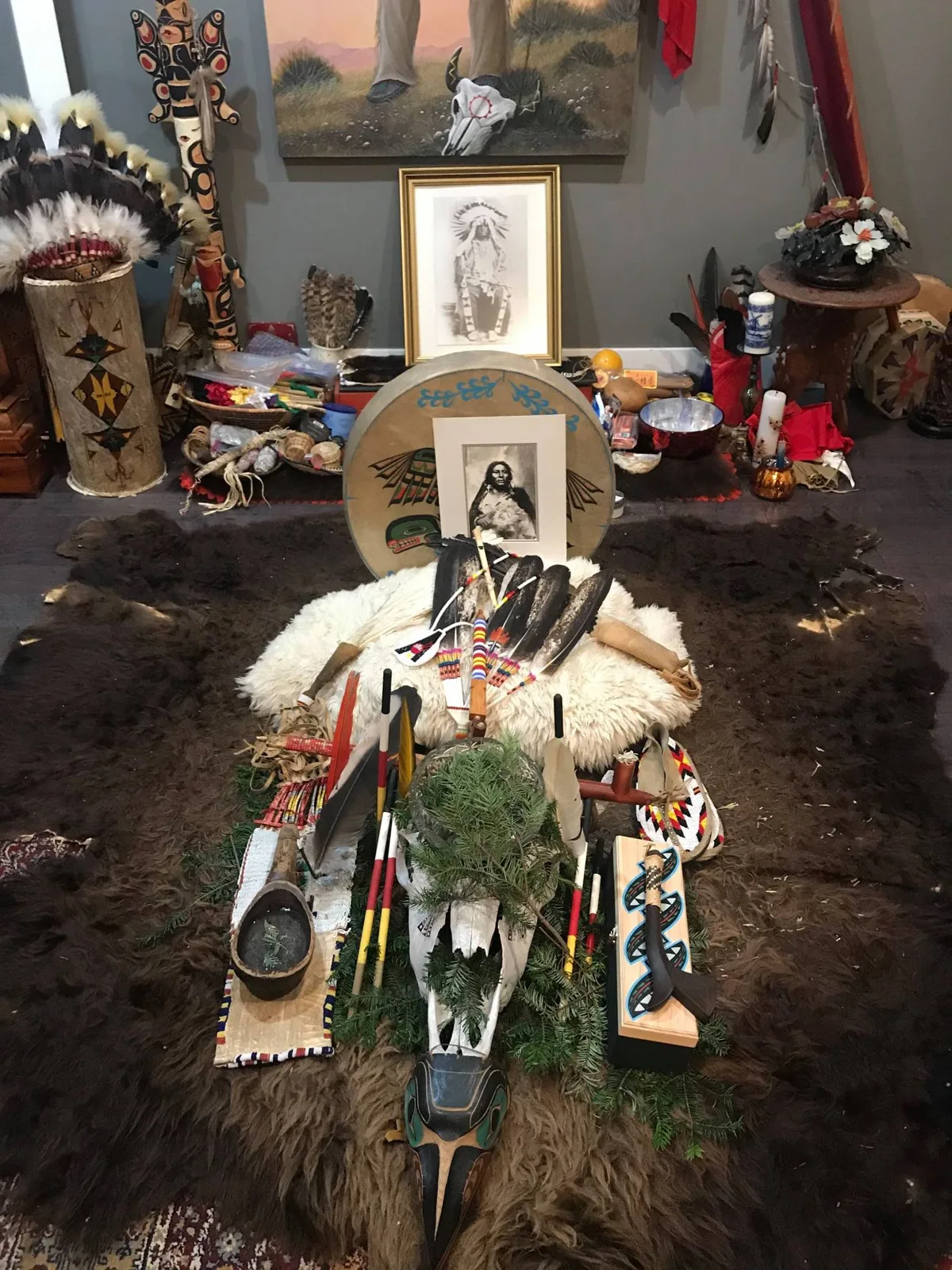 A ceremonial altar on a buffalo robe with eagle feathers, beaded pipe bags, a painted skull with cedar boughs, and a painted drum holding a historic portrait of a Native man.