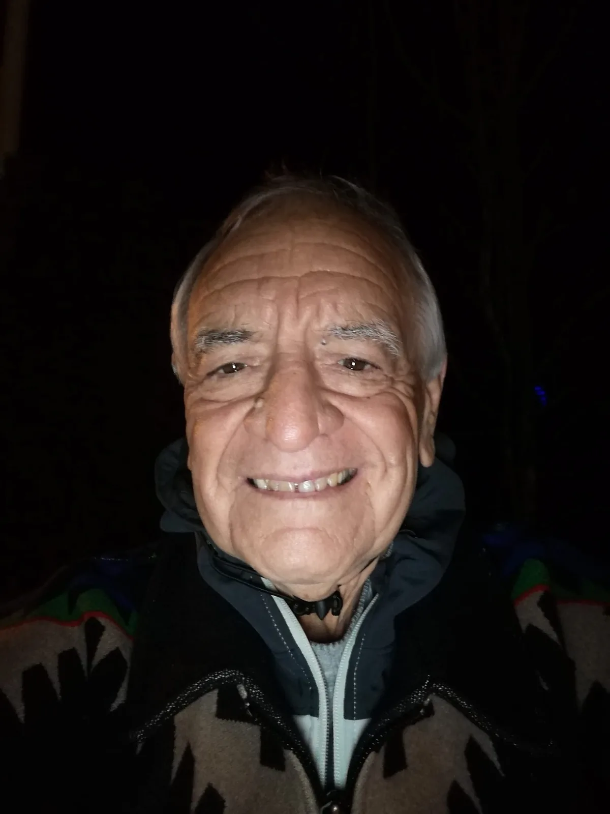 Night-time selfie of a smiling elder with white hair, wearing a dark jacket under a patterned Native-design blanket coat, against a black background.
