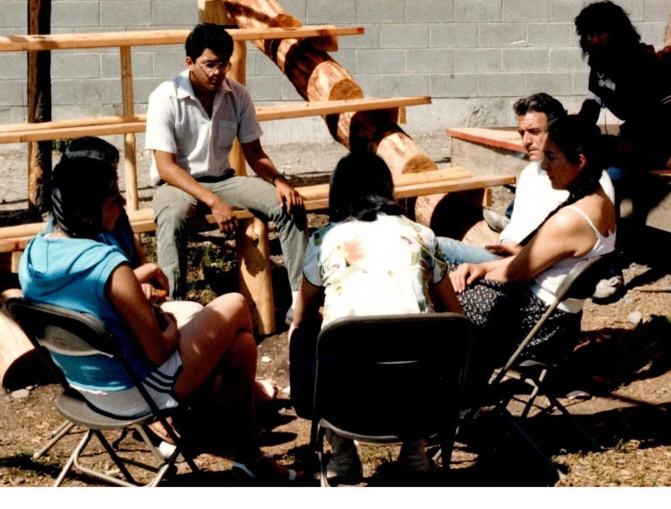 A small group of people sit in a circle on folding chairs and log benches outdoors beside a cinder-block wall, talking together on a sunny day in 1985.