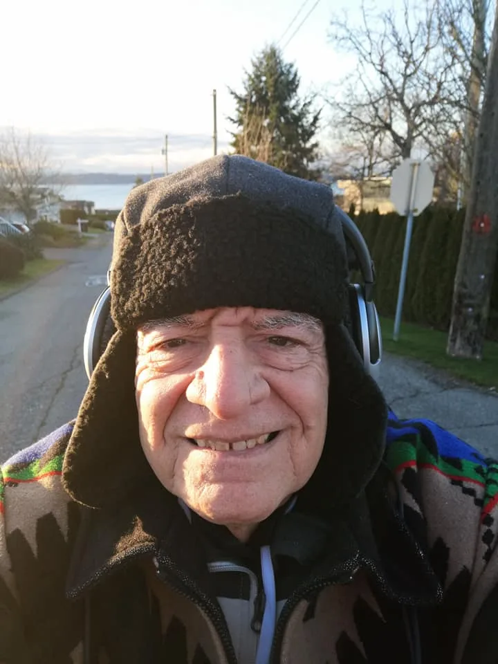 Phil Lane Jr. smiling in a selfie, wearing a black fleece trapper hat, headphones and a patterned jacket, on a residential street sloping down toward the water.
