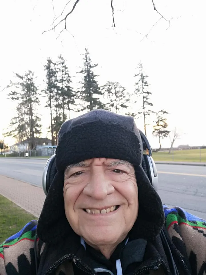 Phil Lane Jr. smiling outdoors in a black fleece hat, headphones and patterned jacket, beside a road lined with tall evergreen trees under a pale sky.