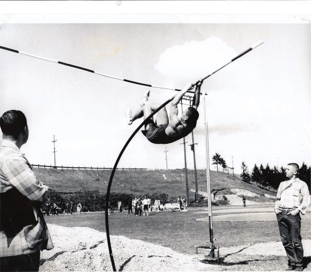 Black-and-white photo of a young athlete arching over a pole vault crossbar on a bending pole, as two men in plaid shirts watch from beside a sand pit on a grassy field.