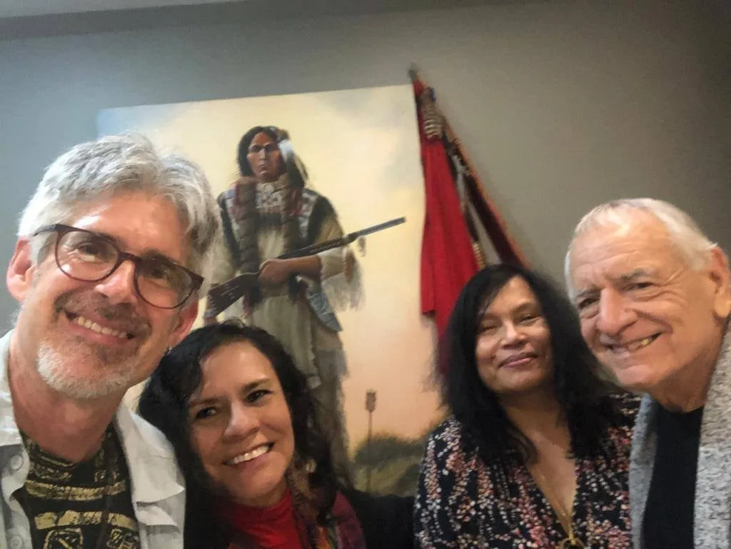Four smiling people pose together for a selfie in front of a painting of a Native warrior holding a rifle, beside a red flag on a staff.