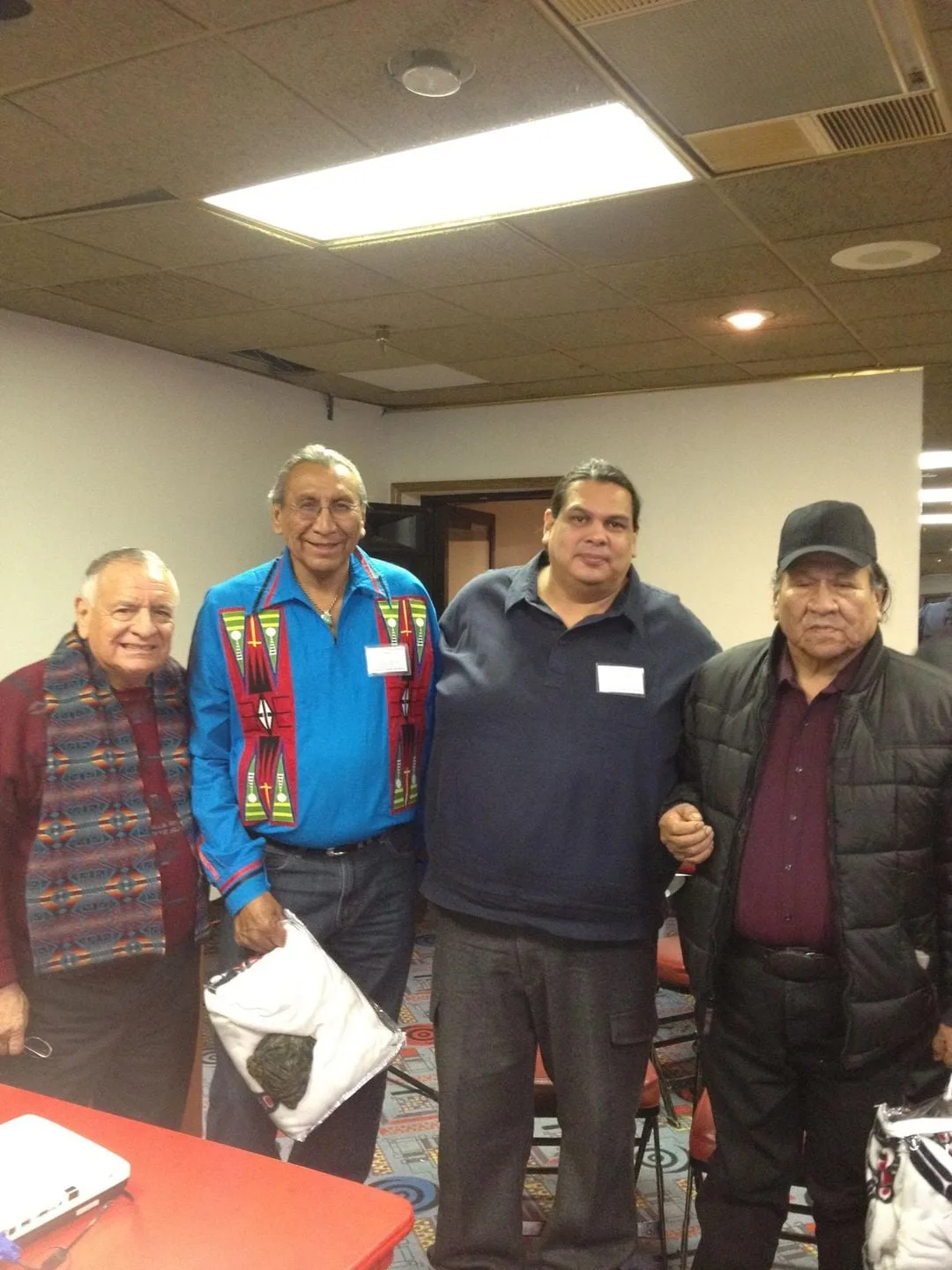 Four Indigenous men stand together smiling in a meeting room; one wears a Pendleton-patterned scarf, one a blue ribbon shirt, one a navy polo, and an elder a black cap and jacket.