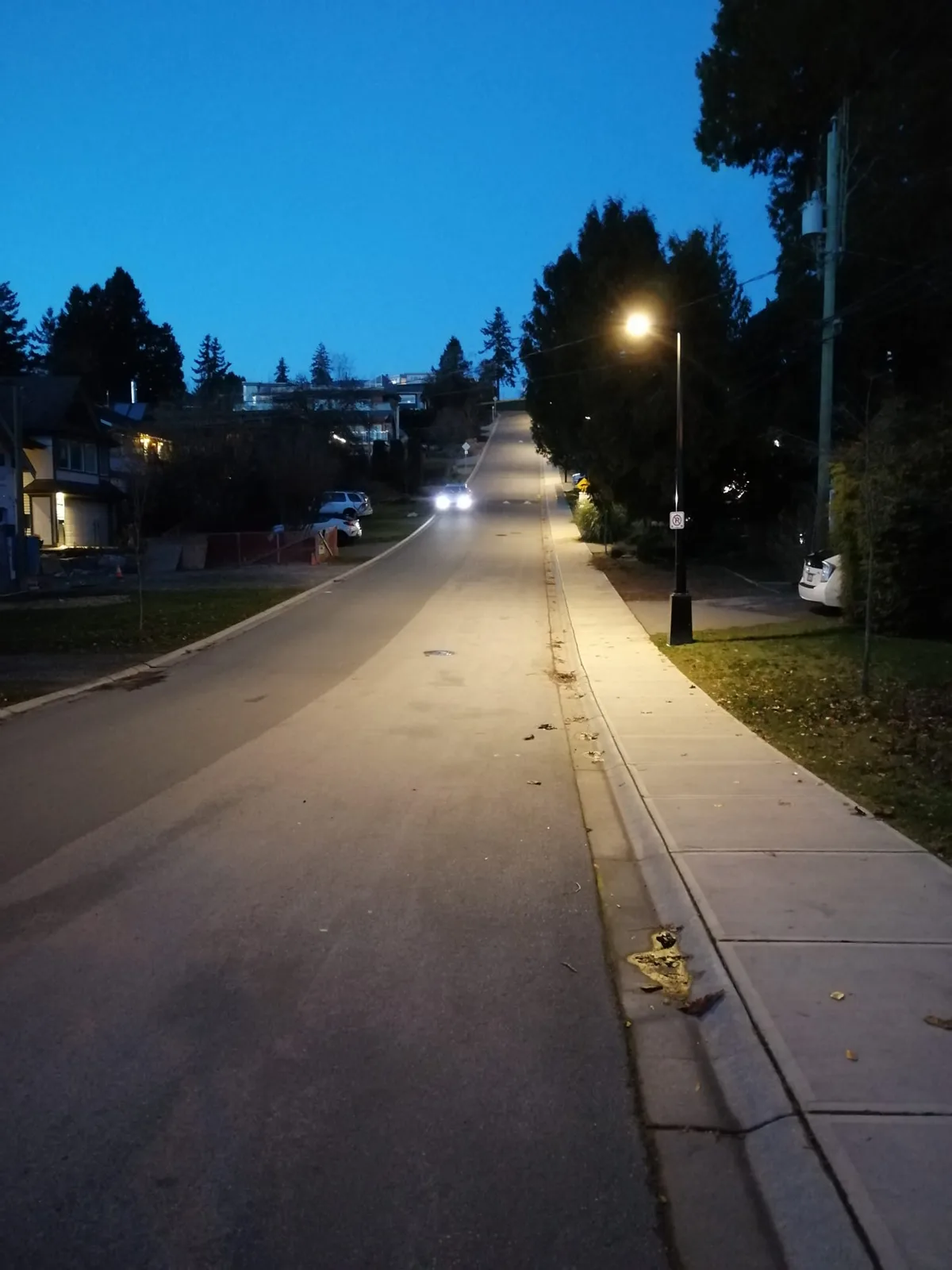 A residential street at dusk climbing steeply uphill, lit by a streetlamp beside the sidewalk, with a car's headlights glowing near the top of the hill.