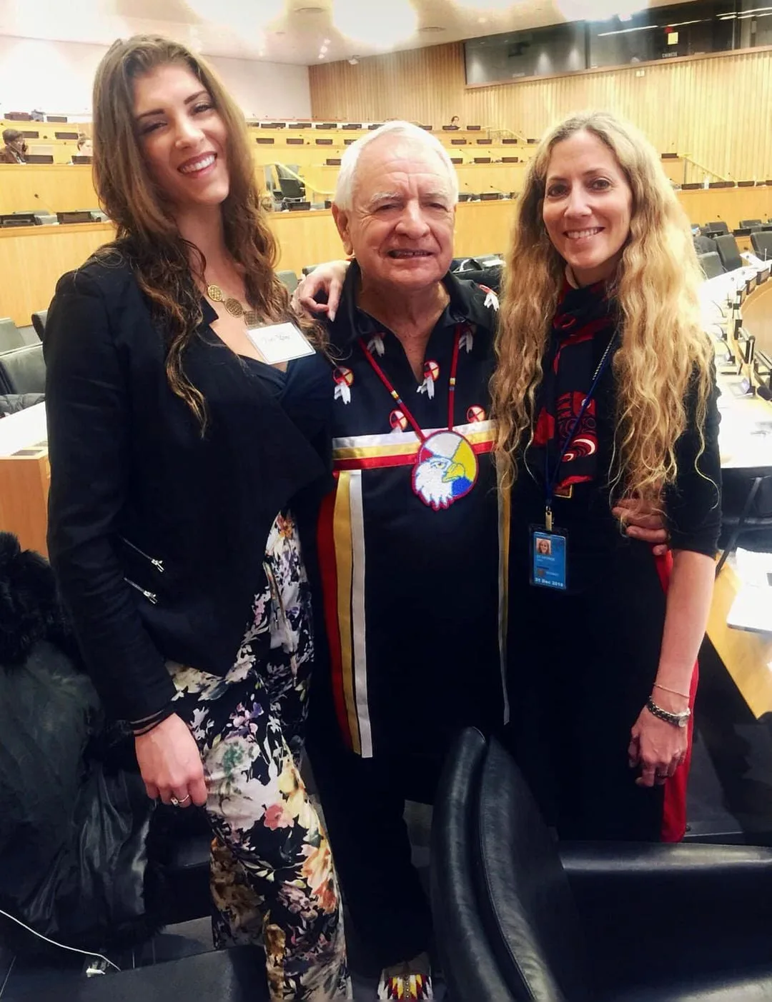 Phil Lane Jr., in a black ribbon shirt with an eagle medallion, stands smiling between Toni Lane Casserly and Chunkshi Shivani in a UN Headquarters conference room.