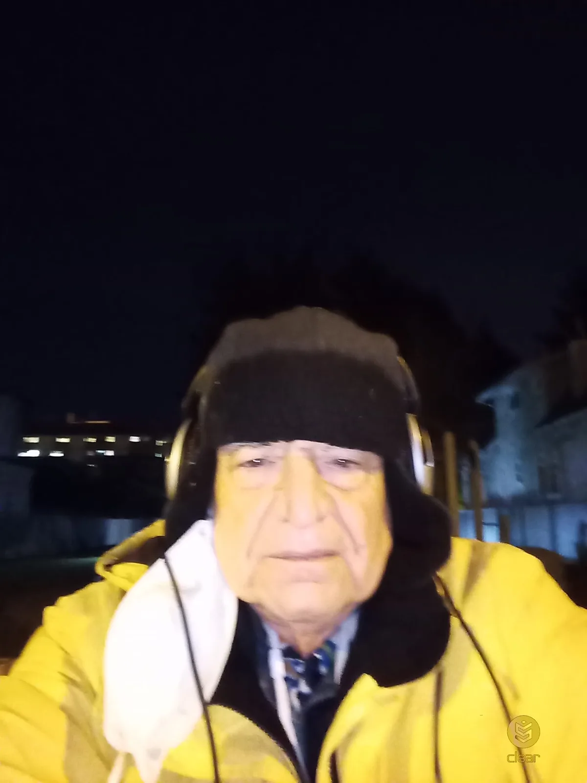 An elder in a black winter hat, headphones and a bright yellow jacket takes a selfie outdoors at night, a face mask hanging from one ear and lit buildings behind him.