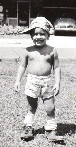 Black-and-white vintage snapshot of a smiling young boy standing shirtless on a lawn in a floppy cap, shorts, rolled socks and boots, with a house and an old car behind him.