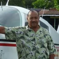 Leonard Gavin, an older man in a green floral shirt, stands in front of a small white airplane with trees and a building behind him.