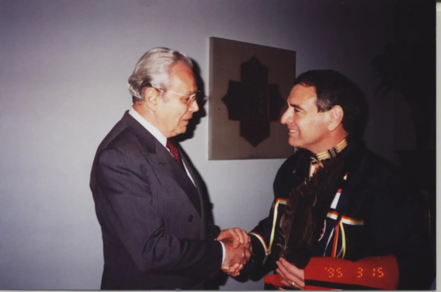 Javier Pérez de Cuellar, a gray-haired man in a pinstripe suit, shakes hands with a smiling man in a dark ribbon shirt holding a feather, beside a wall plaque; dated 3/15/95.