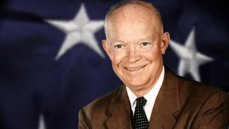 President Dwight D. Eisenhower, smiling, in a brown suit and polka-dot tie, in front of a dark blue background with white stars from an American flag.