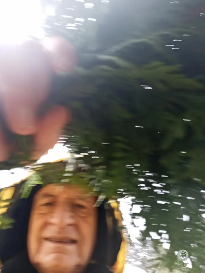 A blurry selfie of an older man in a dark hooded rain jacket, holding cedar boughs overhead as rain falls.