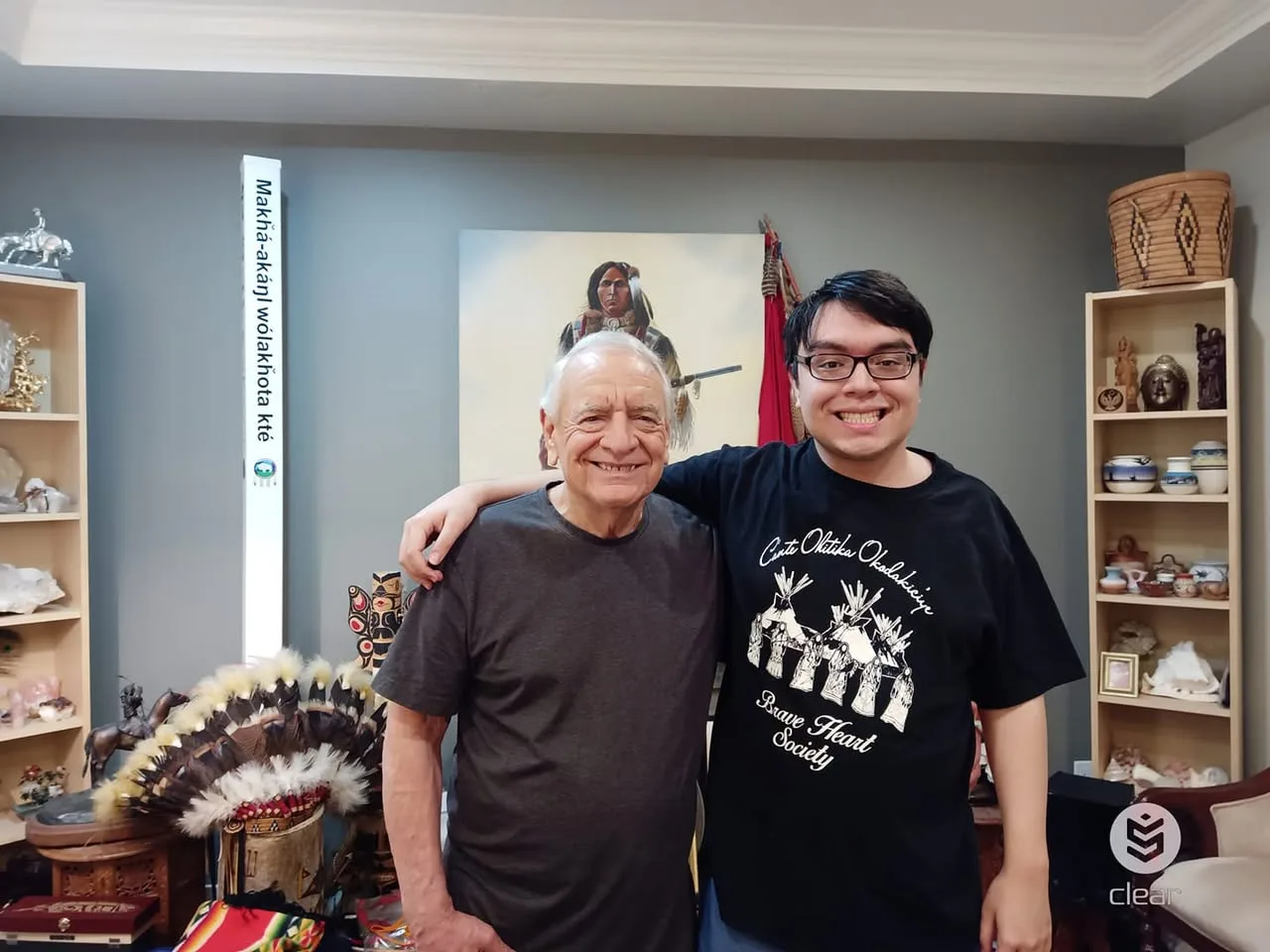 Phil Lane Jr. and his son Tiger Nathan, Mato Gi, smiling arm in arm in a room with a feather headdress, a painting of a Native warrior, and shelves of pottery and baskets.