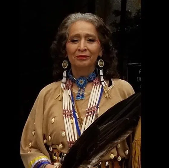 A smiling woman with gray hair wearing a tan buckskin dress with elk teeth, a long bone hairpipe necklace, beaded star medallion and earrings, holding an eagle feather fan.
