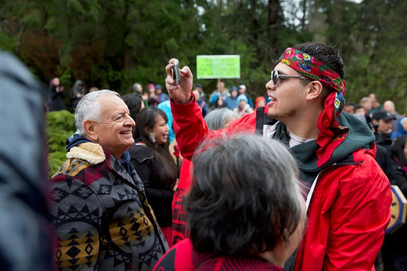 A smiling white-haired elder in a patterned wool coat stands in a crowd outdoors beside a young man in a red jacket and floral headband holding up a phone, with a protest sign behind them.