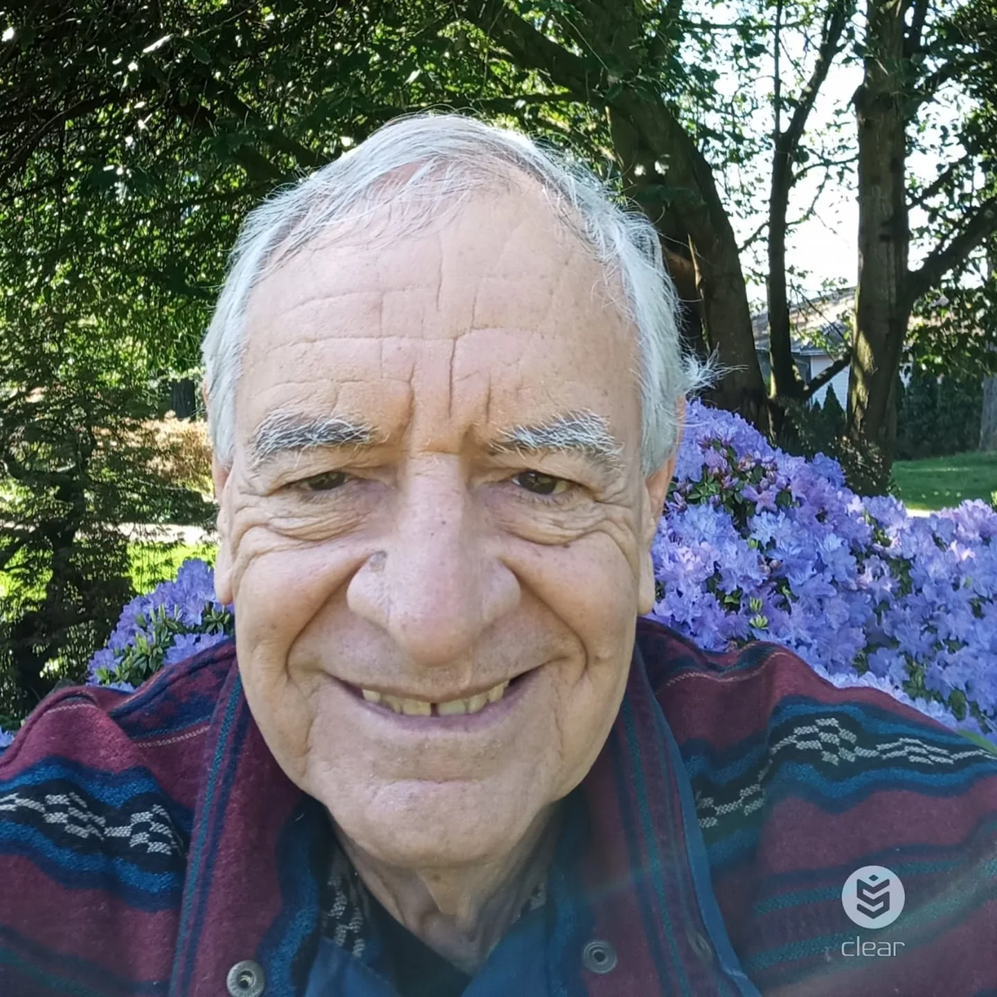 A smiling elder with white hair, wearing a maroon and blue patterned jacket, outdoors in front of purple azalea blossoms and green trees.